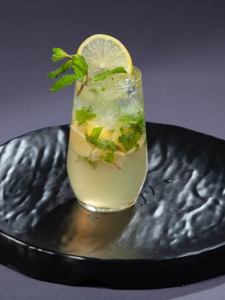 Tall glasses of iced shikanjvi with lemon slices and mint, beside halved lemons and a jug of the drink