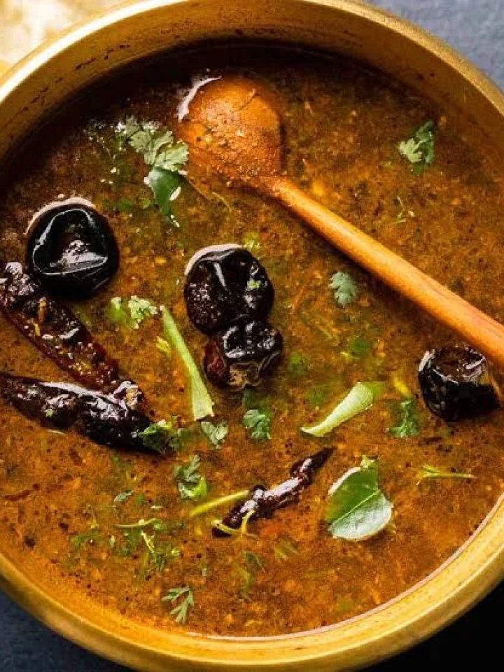 A bowl of red-orange tomato tamarind rasam topped with curry leaves and coriander, served with rice and a papad